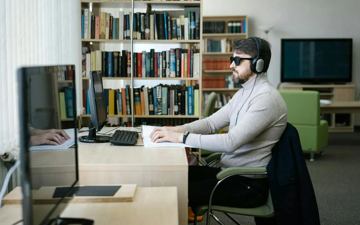 A man wearing dark glasses and headphones sits at a desk with a computer, one hand resting on a page in front of him
