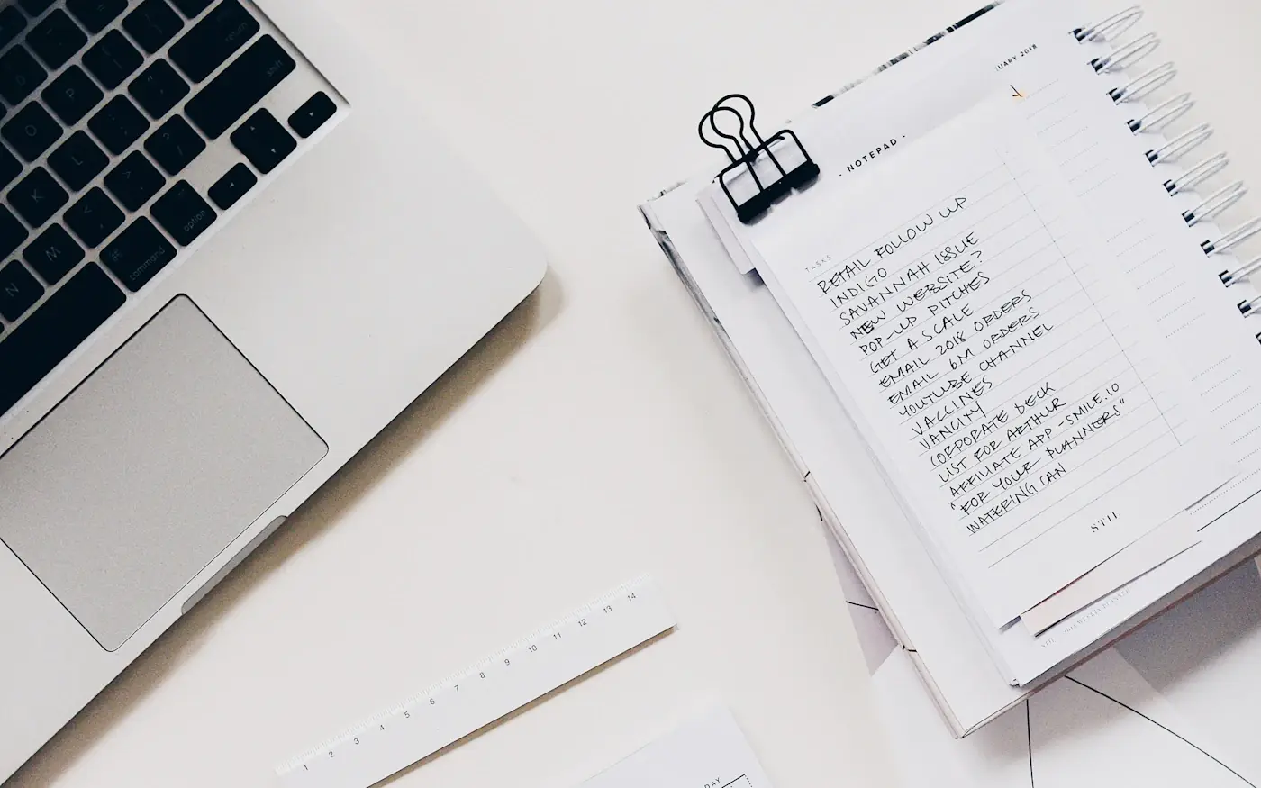 A laptop beside a notebook with a handwritten to-do list on a white desk